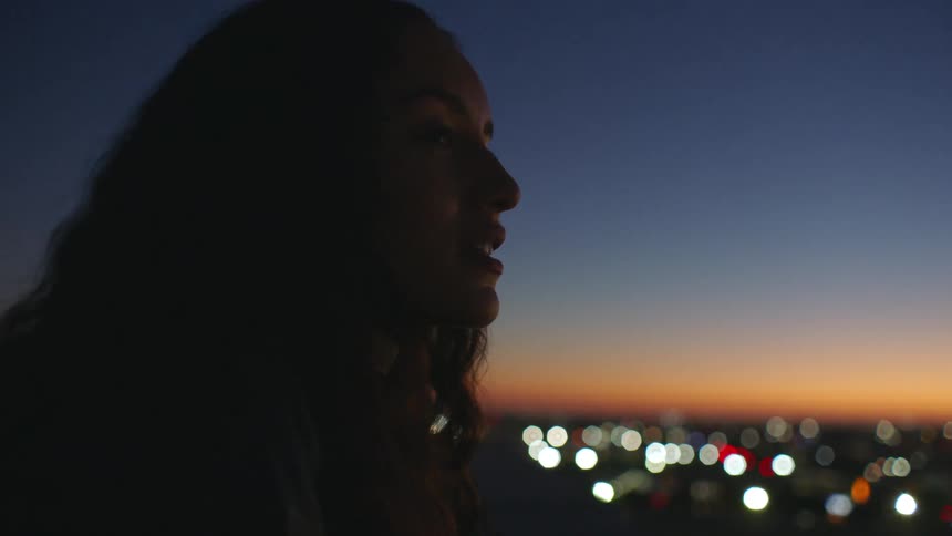 A young woman standing on a rooftop at night smiling as she looks out over the city while the blurred lights glow brightly in the background