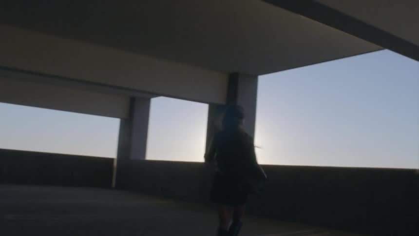 A young woman runs toward the balcony of a parking ramp at sunset smiling as she arrives warm sunlight streaming over her as the sky glows behind her