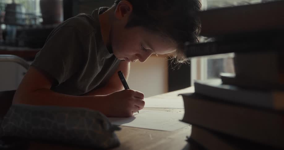 A young boy sits at a wooden table focusing intently as he writes on a piece of paper with a black pen a tall stack of books sits nearby in the dimly lit room with soft light from a window illuminating his work
