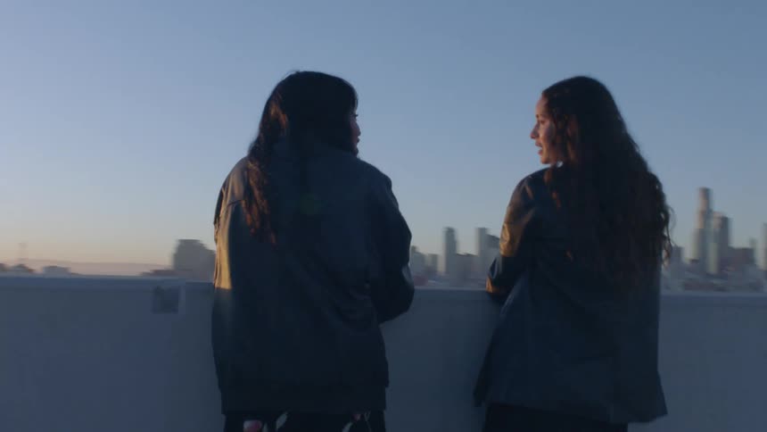 Two women stand on a rooftop overlooking a sprawling city skyline as the sun sets in the distance they lean against a concrete ledge together sharing a quiet moment in the soft golden light of dusk
