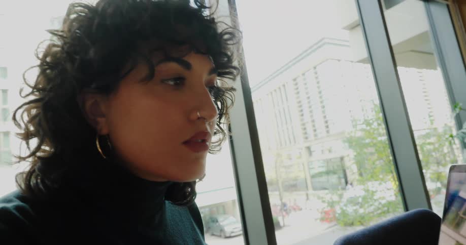 A young woman sitting at a desk beside a window at midday typing on her computer as bright natural light fills the room