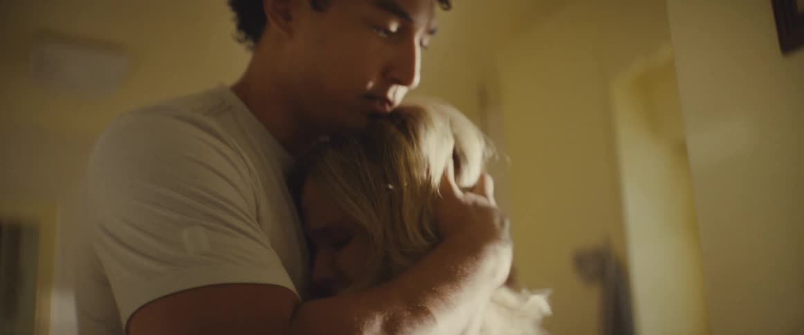 A young man with curly hair looks down and pulls a blonde woman into a close comforting embracethe woman rests her head against his chest as he holds her securely in a dimly lit room