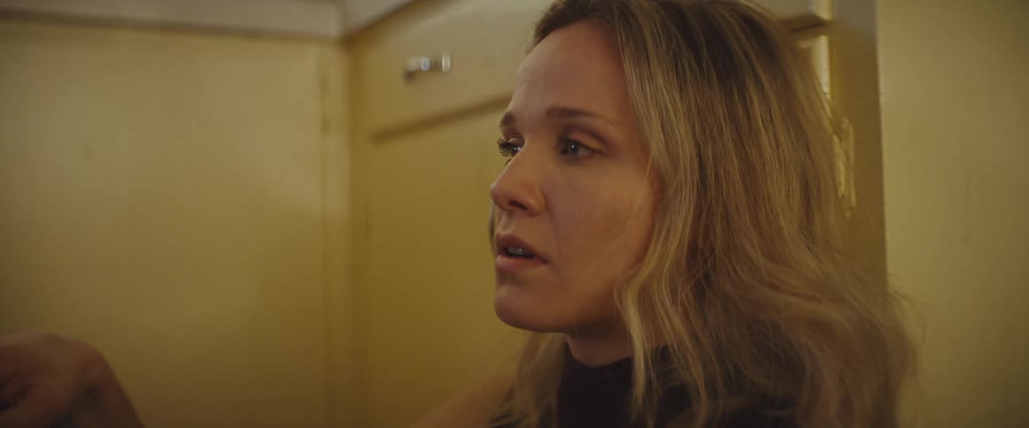 A close view of a blonde woman looking aside with a worried expression while sitting in a kitchen with yellow cabinets