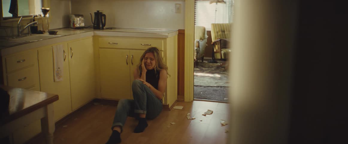 Crying woman slides down to sit against the kitchen cabinets next to broken dishes while a man stomps out after a fight