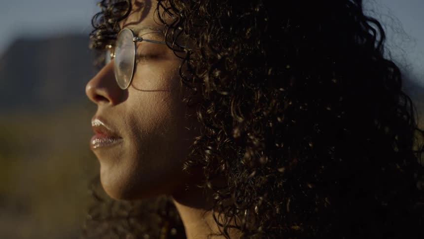 A woman with dark curly hair and glasses is captured in a closeup profile against a sundrenched desert backdrop the golden hour light illuminates her features and the texture of her hair while she remains perfectly still