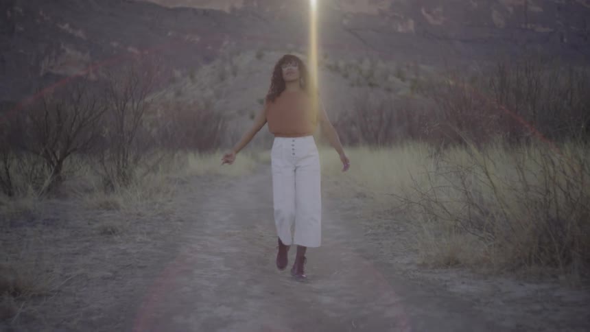 Woman walking on a path cross crossing her legs as she walks with her arms out and a mountain in the distance