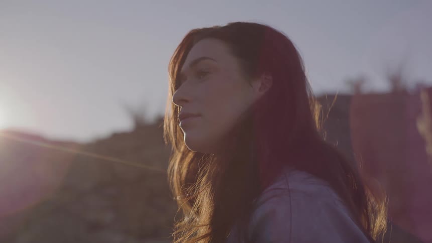 A woman with long dark hair looks out over a desert landscape during a bright sunset with a prominent lens flare as she pushes her hair behind her ear