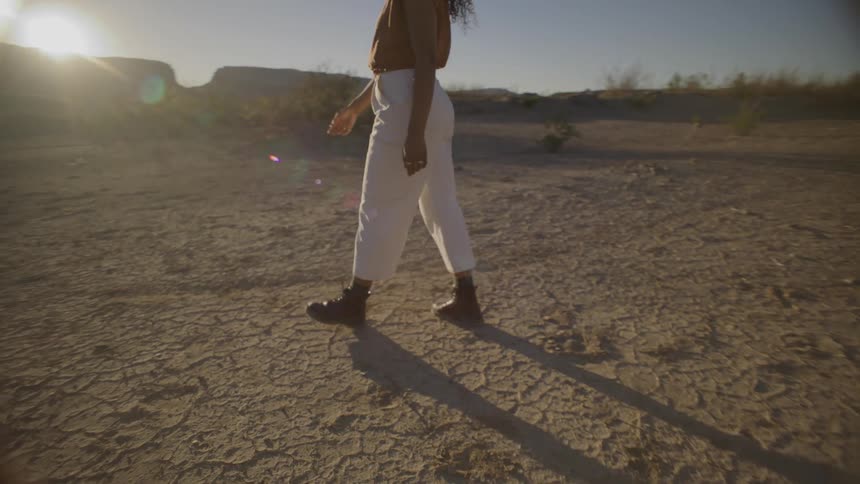 A woman with long curly hair wearing white trousers and dark boots walks across a cracked desert landscape toward the setting sun