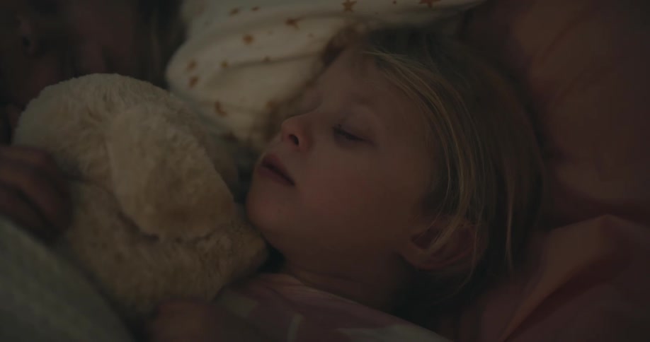 Young girl holding a stuffed animal and yawning in her sleep next to another sleeping young girl on a bed indoors during night time