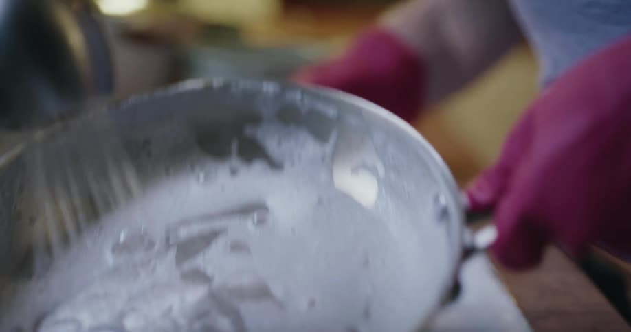 A person wearing red gloves uses a sponge to scrub a soapy stainless steel pan in a kitchen sink after scrubbing they move the pan under a running faucet to rinse away the white suds
