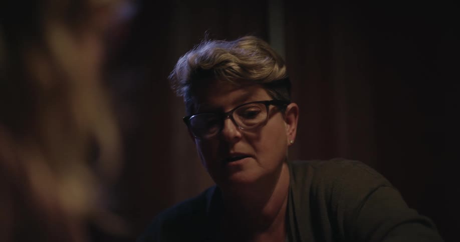 A woman with glasses works with a child to arrange natural items like pinecones and sticks into a small displaythe two sit together in a dimly lit room focusing intently on the craft project as they place their hands on the centerpiece