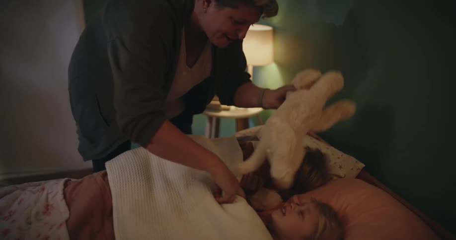 Adult woman putting two female children to sleep by giving them a toy and kissing them in bed indoors during night time