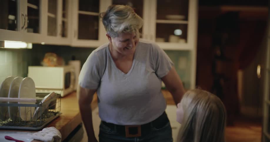 An older woman with short hair celebrates with a young girl in a kitchen giving her a highfive before they walk out of the room together the scene takes place in a warmly lit kitchen featuring white cabinets a wooden countertop and a dish rack by the