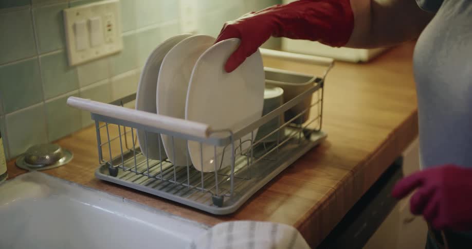 Adult woman wearing rubber gloves and young girl putting dishes on dish drying rack over a counter on a kitchen indoors
