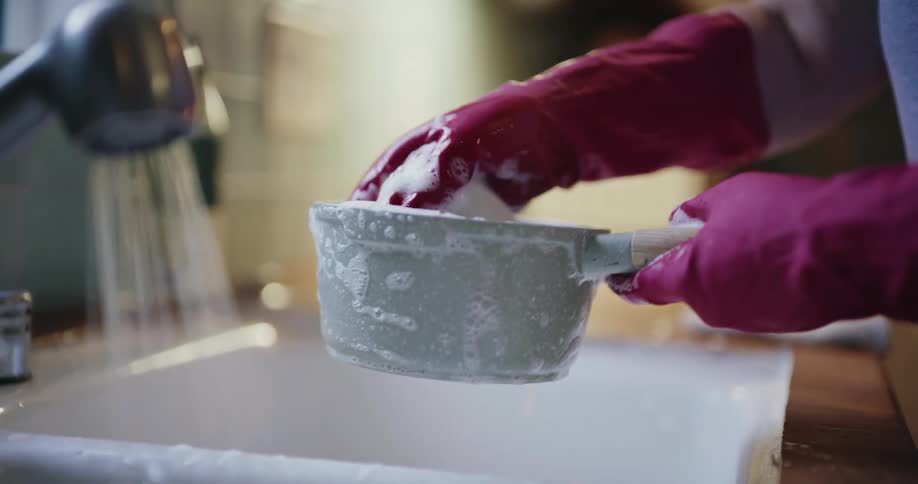 A close view of a woman wearing pink rubber gloves using a soapy sponge to scrub a grey cooking pot in a kitchen sink with water running from the faucet