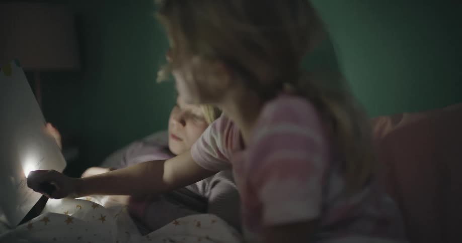 Two young girls sit together in a dimly lit bedroom while using a flashlight to look at a colorful picture book before bed