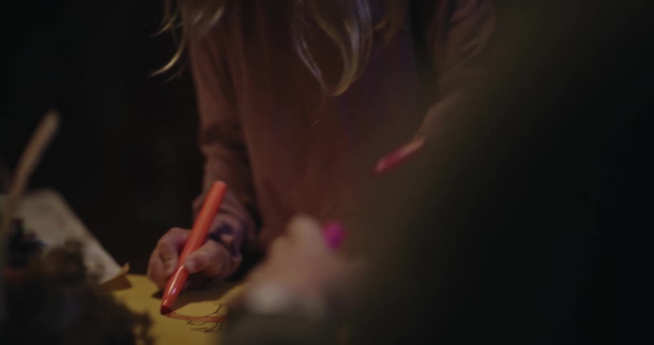 A girl and another person blurred sit at a table using markers to draw pictures and color