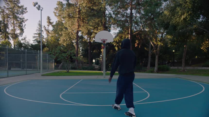 Person performs jump shot on outdoor blue court surrounded by trees