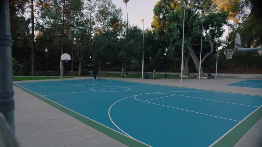 A wide view of a man playing basketball alone on a blue outdoor court surrounded by tall green trees during a quiet afternoon