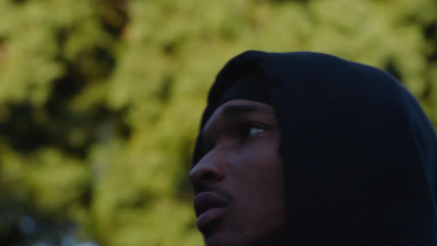 A close view of a man wearing a black hoodie looking up with a focused expression on an outdoor basketball court with green trees in the background