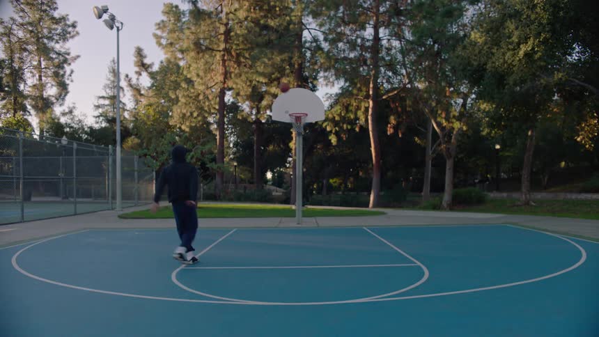 A young man in a dark hoodie practices his jump shot on an outdoor blue basketball court surrounded by trees in a closeup shot he is seen holding the basketball tightly against his chest with a solemn and focused expression