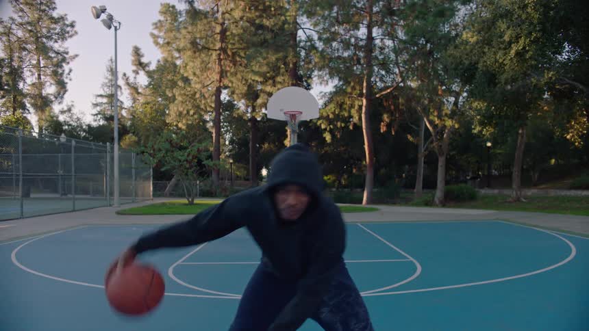 Young adult black man dribbling a basketball between his legs and walking at a sports court in a park outdoors during day time