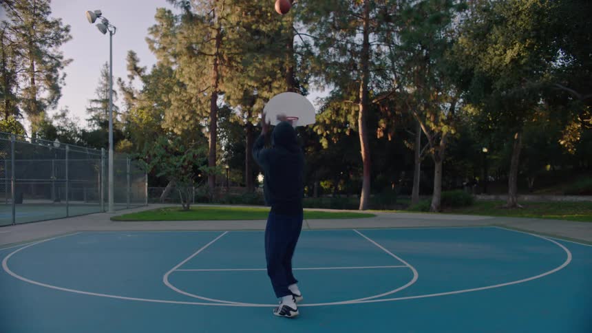 A young man wearing a hoodie playing basketball on an outdoor court at sunset warm light stretching across the pavement