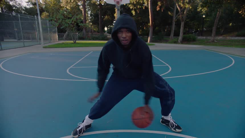 Young black adult man dribbling a basketball between his legs at a sports court on a park outdoors during dusk or dawn