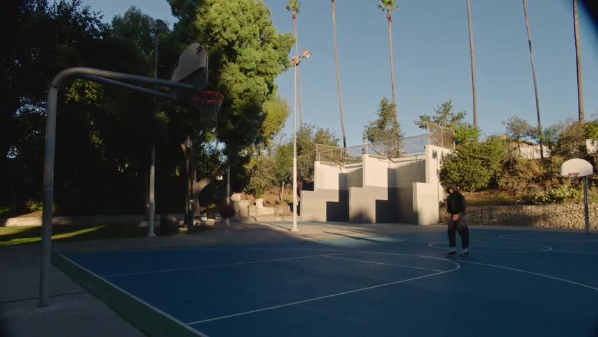 A man in a black hooded sweatshirt and dark trousers shoots a basketball on an outdoor blue court surrounded by tall palm trees and lush greenery under a clear sky