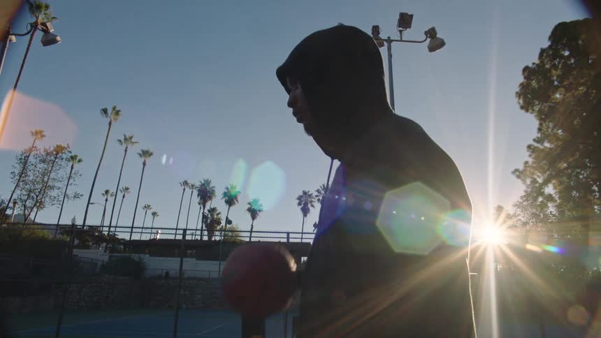 A man wearing a dark hoodie dribbles a basketball on an outdoor court during a sunny morning captured from a low angle that highlights the surrounding palm trees and stadium lights against a bright sky with visible lens flares