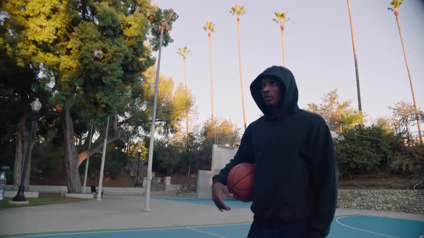 A young man in a black hoodie walks across an outdoor blue basketball court at dusk dribbling a ball while the camera follows him in a lowangle tracking shot past palm trees and park greenery