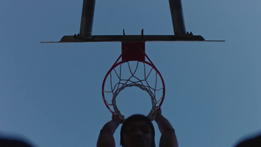 A low angle view looking up at a basketball hoop as a man jumps and performs a slam dunk against a clear blue sky