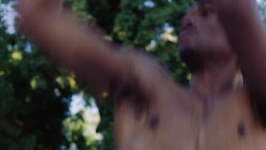A young black man shirtless throwing a basketball toward the hoop on an outdoor court at midday under bright sunlight