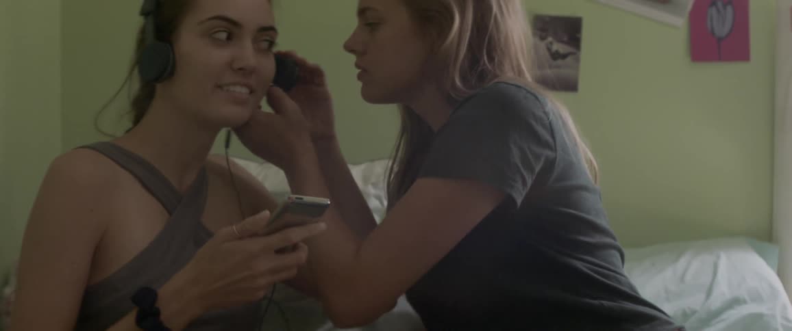 Two women sit on a bed together and listen to music with headphones