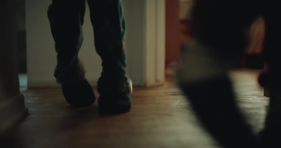 A person wearing blue socks and slippers walks across a polished wooden floor past a doorway another person follows closely behind as they move through the warmly lit hallway of a home