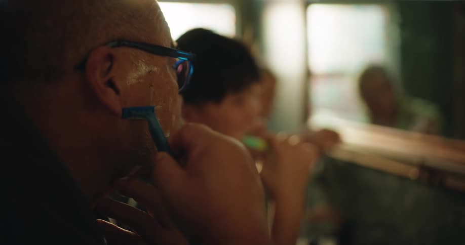 Man shaves in front of a mirror while a child brushes teeth in the background