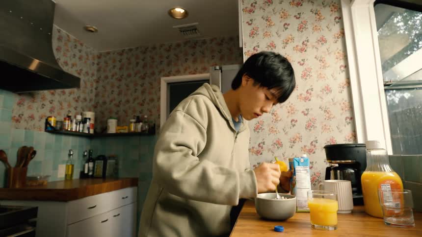 A young man wearing a grey hoodie stands in a kitchen with floral wallpaper and pours milk into a bowl of cereal before eating and drinking orange juice from a glass