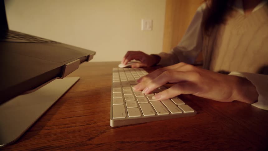 Hands typing on a keyboard on a wooden desk beside a laptop indoors