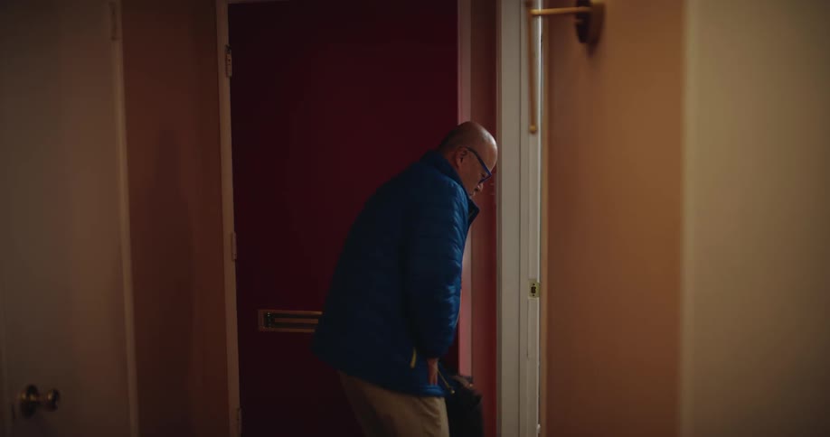 After walking down a hallway and opening a red door a man stops to frantically search his jacket pockets before turning around and walking back in the opposite direction