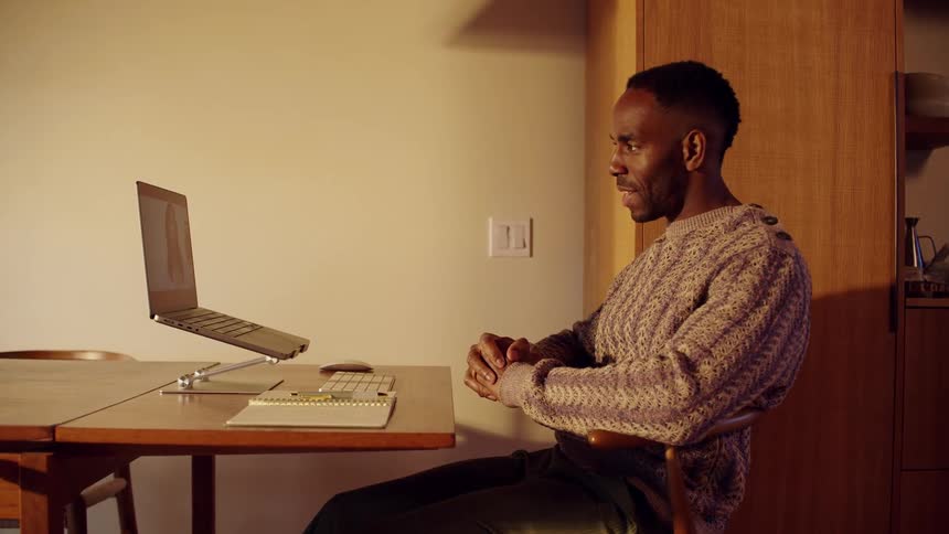 Man speaks during a video call while seated at a table with a laptop raised on a stand