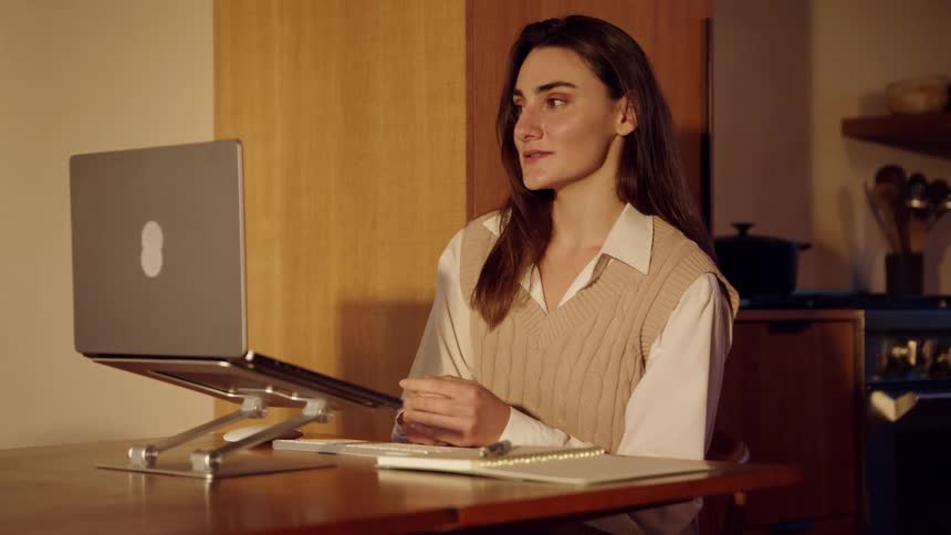 A woman with long brown hair wearing a beige sweater vest over a white shirt sits at a wooden table and talks during a video call on her laptop in a warm room at night