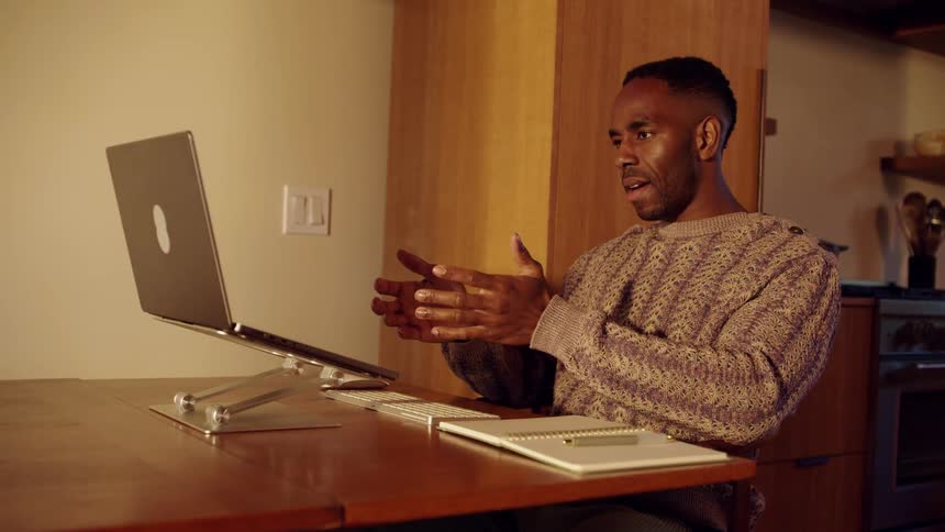 Man sitting at a table near the kitchen gestures during a video call on a laptop