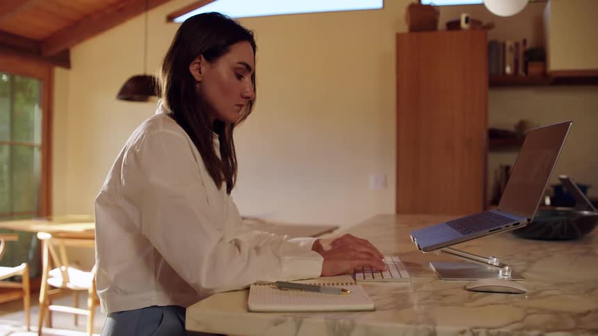 A woman wearing a white long sleeve shirt and blue trousers sits at a marble kitchen counter and types on a keyboard with a laptop on a silver stand in a home with wooden ceilings