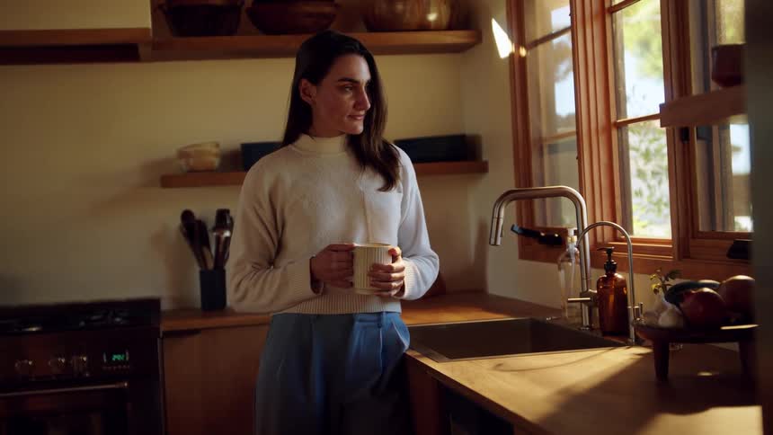 A young woman wearing a white turtleneck sweater and blue trousers stands in a rustic kitchen while holding a ceramic mug and looking out of a window as morning sunlight illuminates the room