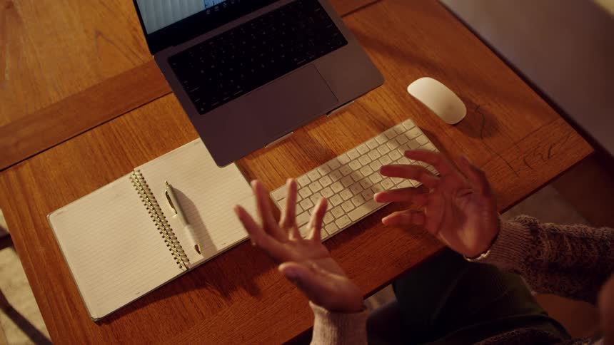 Person gestures in front of open laptop on wooden desk with notebook and keyboard beside