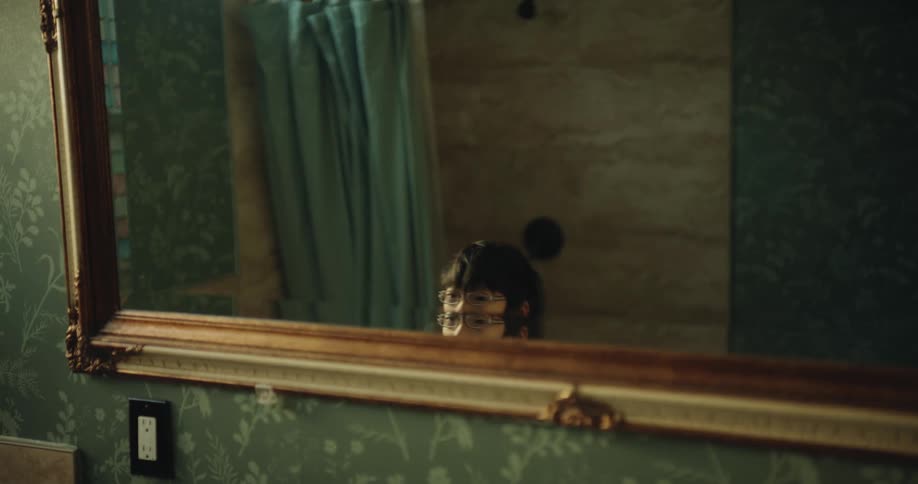 In this short video a young child wearing glasses is seen in a bathroom standing in front of a mirror with an ornate gold frame the child playfully ducks up and down out of the reflections view eventually raising their arms in a celebratory gesture b