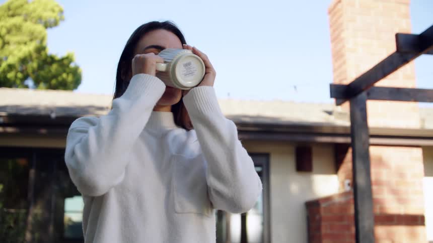 A young woman drinks from a coffee mug while standing outside her house