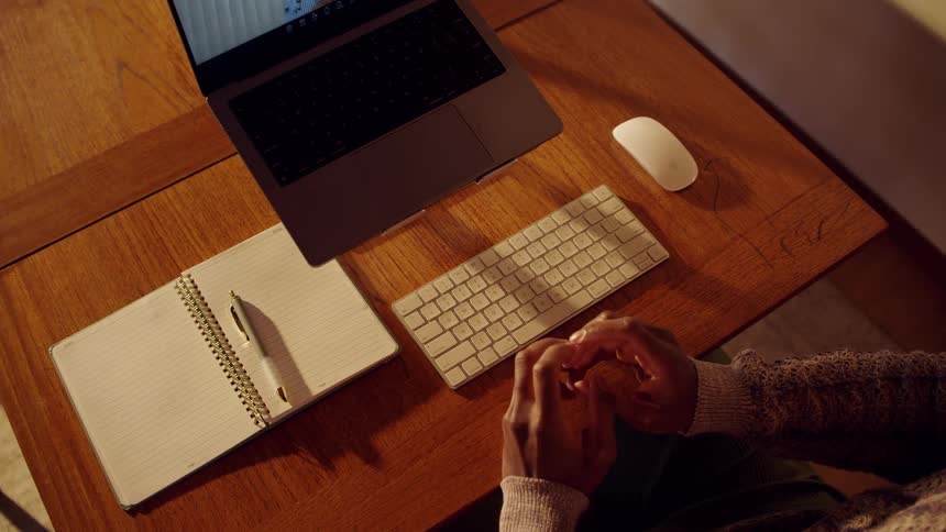 Person seated at desk with open notebook laptop and clasped hands in welllit setting
