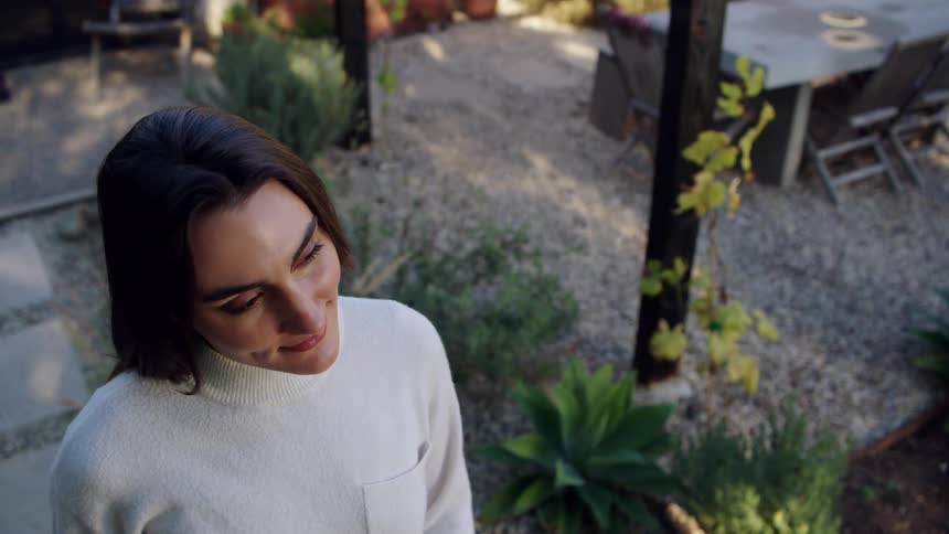 From a high angle a woman with a serene expression looks down and smiles gently while standing in a peaceful landscaped backyard
