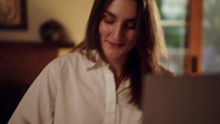 A woman with long dark hair wearing a white button down shirt looks at a computer screen and smiles before writing in a notebook with a gold pen at a desk